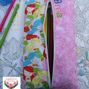 Puede incluir: Estuche rectangular con un diseño de sirenas y un estampado floral rosa. El estuche está parcialmente abierto, revelando lápices de colores. La palabra "Brea" está bordada en el lado rosa. Debajo hay un libro para colorear.
