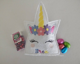 Puede incluir: Bolsa de fieltro blanco en forma de unicornio con una corona de flores rosa, amarilla y morada, pestañas bordadas y el nombre "Brea" cosido en la parte delantera. La bolsa está llena de caramelos Tic Tac y pequeños huevos de chocolate.