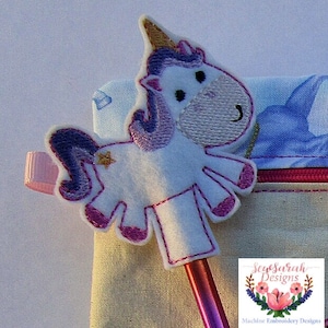 May include: A white felt unicorn pencil topper with purple and gold accents. The unicorn is holding a star in its mouth. The topper is on a pink and blue fabric background.