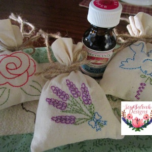 May include: Three small white fabric bags with embroidered designs of a rose, lavender, and a bird. The bags are tied with twine and are sitting on a green and white fabric background. A small bottle of essential oil is in the background.