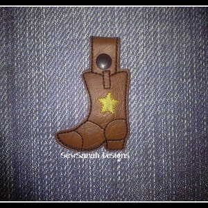 May include: Brown leather boot keychain with a gold star and black stitching. The keychain is on a blue denim background. SewSarah Designs.