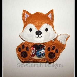 May include: A felt fox-shaped candy holder with a white face and brown ears, paws, and tail. The fox is sitting with its paws outstretched and has a small opening in its belly to hold candy. The fox is holding a variety of small candies, including M&Ms and Reese's Pieces. The text "SewSarah Designs" is printed below the fox.