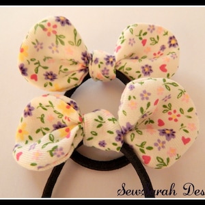 May include: Two white fabric hair scrunchies with a floral print in shades of pink, yellow, purple, and green. The scrunchies are tied into bows.