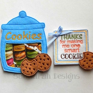 May include: Blue felt cookie jar with a window showing colorful macarons. The jar has the word "Cookies" embroidered on the front. There are two brown felt cookies next to the jar. A blue felt tag with the text "THANKS for making me one smart COOKIE" is attached to the jar.