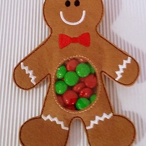 May include: A gingerbread man-shaped felt ornament with a clear plastic window in the center. The window is filled with red and green candy. The gingerbread man has a white smile, black eyes, and a red bow tie.