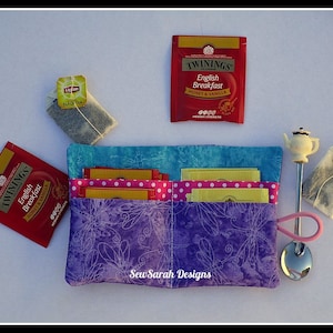 Peut inclure: Un porte-thé en tissu violet et bleu avec un motif floral. Le porte-thé a quatre poches pour les sachets de thé et une petite poche pour une cuillère à thé. Le porte-thé est étiqueté "Sew Sarah Designs".