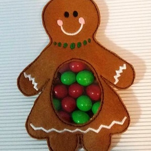 May include: A felt gingerbread person with a clear plastic window in the middle. The window is filled with red and green candy. SewSarah Designs is written at the bottom of the image.