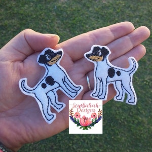 May include: Two white and black embroidered patches of a dog with black spots. The dog has a black nose and brown eyes. The patches are on a hand with a pink bracelet.