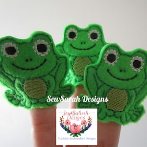 May include: Three green felt finger puppets shaped like frogs with black eyes and white pupils. The puppets are on fingers and the text "Sew Sarah Designs" is visible below.