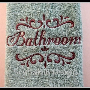 Puede incluir: Una toalla de color verde claro con la palabra "Bathroom" bordada en letras cursivas marrones. La palabra está enmarcada por dos remolinos decorativos.