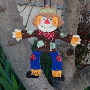 May include: A felt scarecrow with a brown hat, orange hair, a red bandana, and blue jeans. The scarecrow has a brown felt body with green and brown patches and is decorated with gold buttons and cactus patches.