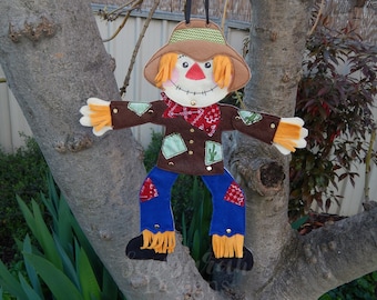 May include: A felt scarecrow with a brown hat, orange hair, a red bandana, and blue jeans. The scarecrow has a brown felt body with green and brown patches and is decorated with gold buttons and cactus patches.