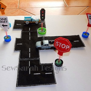 May include: A felt play set for children featuring a black road with white lines, a traffic light, a stop sign, a give way sign, a one way sign, and two toy cars.