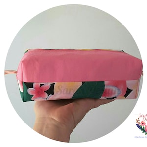 Puede incluir: Una bolsa con cremallera rosa con estampado floral y una solapa superior rosa. La bolsa se sostiene en una mano.