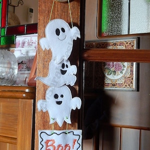 May include: Three white felt ghosts with black eyes and mouths are hanging from a door with a white felt sign that says "Boo!" SewSarah Designs is written below the sign.