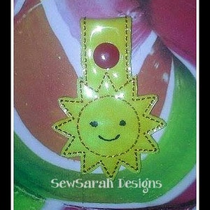 May include: Yellow felt sun with a smiling face, sewn onto a yellow and red felt key chain. The key chain has a red button in the center.