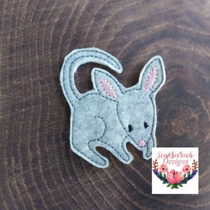 May include: A gray felt appliqué of a small, cartoonish kangaroo with pink ears. The appliqué is on a brown wooden background. SewSarah Designs is printed on a white tag with a floral design.
