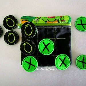 Può includere: Una borsa in tessuto nero con una chiusura a zip e un design delle Tartarughe Ninja. La borsa è piena di cerchi di feltro verde e nero con una X o una O ricamata su di essi. La borsa è per giocare a un gioco di tris.