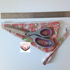 May include: A pink floral zippered pouch with a white zipper and a matching strap. A pair of silver scissors with purple handles are inside the pouch. The pouch is laying on a white surface with a ruler next to it.