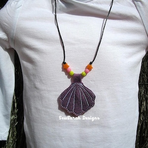May include: A purple felt seashell pendant necklace with a black cord and colorful beads. The necklace is worn by a child. The pendant is decorated with white stitching. The text "SewSarah Designs" is visible at the bottom of the image.
