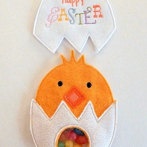 May include: A felt Easter chick with a clear plastic window for candy. The chick is orange with black eyes and a diamond-shaped beak. The chick is in a white felt egg with a white felt top that says "Happy Easter" in colorful letters.