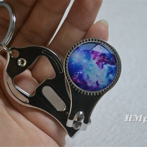 Könnte beinhalten: Ein silberfarbener Schlüsselanhänger mit Flaschenöffner und Nagelknipser. Der Schlüsselanhänger hat ein rundes Design mit Galaxiemotiv in Blau, Lila und Weiß. Der Text "HMpisces" ist sichtbar.