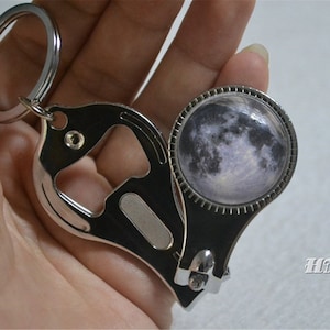 May include: A silver-colored keychain with a bottle opener and a nail clipper. The keychain features a circular design with a detailed image of the moon. The text "HMpisces" is visible in the image.