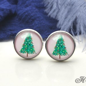 Puede incluir: Un par de pendientes de botón redondos plateados. Cada pendiente presenta un diseño de árbol de Navidad con agujas verdes, adornos coloridos y un tronco marrón, sobre un fondo rosa claro. Los pendientes están sobre una superficie blanca.