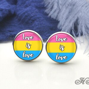 May include: A pair of round, silver-toned earrings. Each earring features a design with pink, yellow, and blue stripes, and the words "Love is love" in white script. The earrings are set against a white surface.