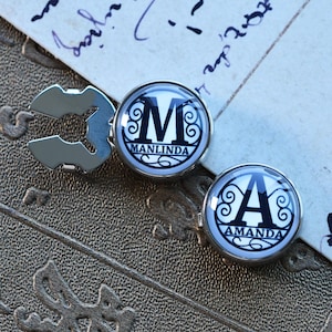 Puede incluir: Dos gemelos de plata con diseños en blanco y negro. Los gemelos presentan las letras "M" y "A" con los nombres "Manlinda" y "Amanda" escritos debajo.