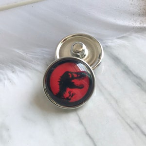 Può includere: Due bottoni a pressione argentati. Uno presenta una silhouette di dinosauro nera su sfondo rosso, con bordo argentato. L'altro bottone è argentato liscio. I bottoni sono su una superficie di marmo bianco e grigio.