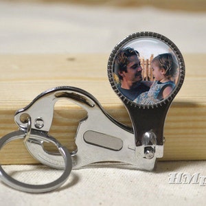 Könnte beinhalten: Ein silberfarbener Schlüsselanhänger mit einem runden Anhänger, der ein Foto von einem Mann und einem Kind enthält. Der Schlüsselanhänger hat einen Flaschenöffner und einen Schlüsselring. Auf hellem Holz.
