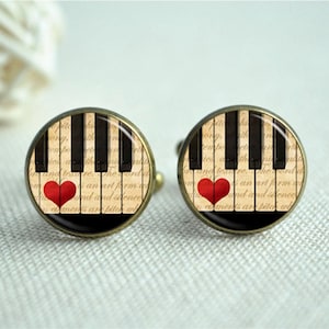 Può includere: Un paio di gemelli con un design a tastiera di pianoforte e un cuore rosso su ciascuno. Lo sfondo è un foglio di musica dall'aspetto vintage.