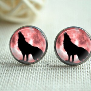 May include: A pair of round stud earrings. Each earring features a black silhouette of a howling wolf against a red and pink background, set in a silver-tone metal. The earrings are displayed on a textured, off-white surface.