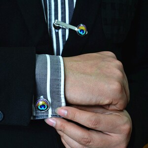 May include: A black blazer with a striped shirt underneath. The shirt has a silver tie bar with a peacock eye design. The cufflinks also feature a peacock eye design.