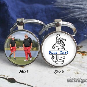 Op de afbeelding: Een ronde, zilveren sleutelhanger met twee zijden. Eén zijde toont een foto van twee golfers op een green. De andere zijde heeft een golfbag-afbeelding met de tekst "Your Text" in blauw. Een golf-thema accessoire.
