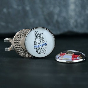 May include: A silver golf ball marker with a white background and a black and white image of a golf bag with golf clubs and the text "Jackson". The marker is attached to a silver clip.