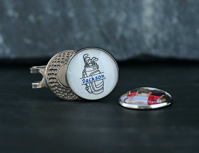 May include: A silver golf ball marker with a white background and a black and white image of a golf bag with golf clubs and the text "Jackson". The marker is attached to a silver clip.