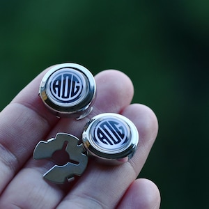 May include: Silver cufflinks with a black and white monogram design. The cufflinks have a circular shape with a shiny, reflective surface. A silver metal piece is also visible.
