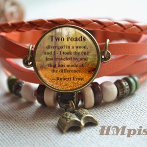 May include: A multi-strand orange leather bracelet with a round charm featuring a quote from Robert Frost's poem "The Road Not Taken". The charm has a picture of a path through a forest with autumn leaves. The bracelet also has wooden beads and a small book charm.