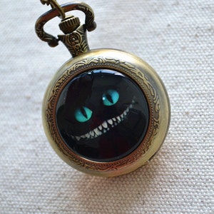 May include: Antique brass pocket watch pendant featuring the Cheshire Cat from Alice in Wonderland. The watch face has a black background with the cat's grinning face, including bright teal eyes and white teeth. The watch is suspended from a matching chain.