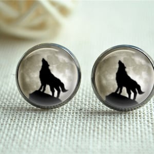 May include: A pair of round stud earrings with a silver-colored setting. Each earring features a black silhouette of a howling wolf against a full moon backdrop. The earrings are displayed on a textured, off-white surface.
