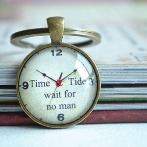 May include: A round, antique gold-toned keychain with a glass clock face. The clock face has black hands and black Roman numerals. The clock face also has the text "Time & Tide wait for no man".