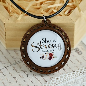 Può includere: Una collana con pendente rotondo in legno con le parole "She is Strong" e "Proverbi 31:25" stampate su sfondo bianco. Il pendente ha un bordo marrone con intagli a forma di cuore e un cordino nero.