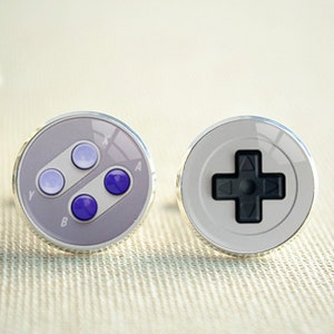 Peut inclure: Paire de boutons de manchette argentés avec un design de manette de jeu vidéo. L'un a des boutons violets et l'autre une croix directionnelle noire, tous deux sur fond blanc et gris.