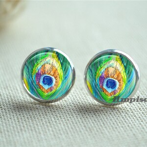 Puede incluir: Un par de pendientes de botón redondos con un diseño de pluma de pavo real. Los pendientes tienen un engaste de metal plateado y una superficie similar al vidrio. El diseño de la pluma presenta tonos de verde, azul, naranja y amarillo.
