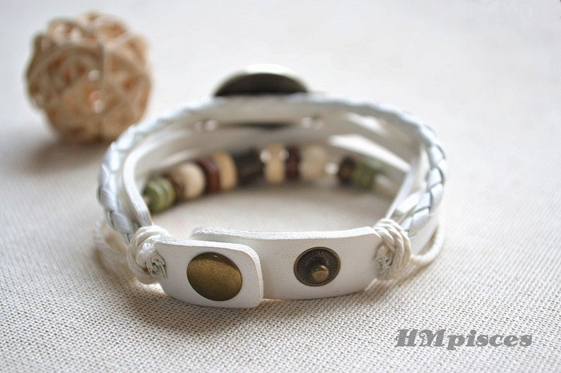 May include: White leather bracelet with a braided design and brown and white beads. The bracelet has a metal clasp.