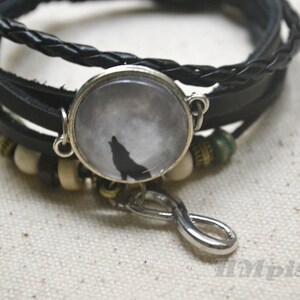 May include: A black leather bracelet with a silver charm featuring a wolf howling at the moon. The charm is a glass dome with a white background and a black silhouette of a wolf. The bracelet also has a silver infinity symbol charm.