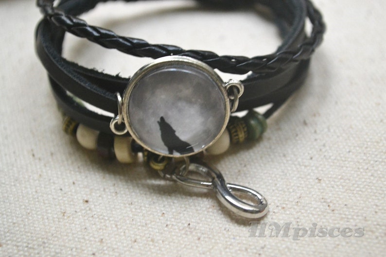 May include: A black leather bracelet with a silver charm featuring a wolf howling at the moon. The charm is a glass dome with a white background and a black silhouette of a wolf. The bracelet also has a silver infinity symbol charm.