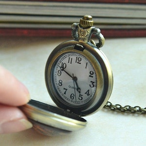 K&ouml;nnte beinhalten: Eine antike Taschenuhr aus Bronze mit rundem Geh&auml;use und passender Metallkette. Das wei&szlig;e Zifferblatt zeigt schwarze arabische Ziffern von 1 bis 12, einen inneren Ring mit Zahlen von 13 bis 24, schwarze Zeiger und das Wort QUARTZ.